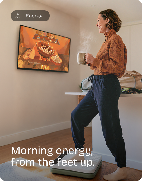 Smiling woman holds a coffee mug while standing on the Kiwii Move board in her kitchen, a game on the TV. Energy: Morning energy, from the feet up.