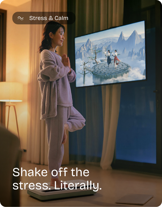 Woman holds a tree pose on the Kiwii Move vibration plate in a lamp-lit living room, a game on the TV. Stress & Calm: Shake off the stress. Literally.